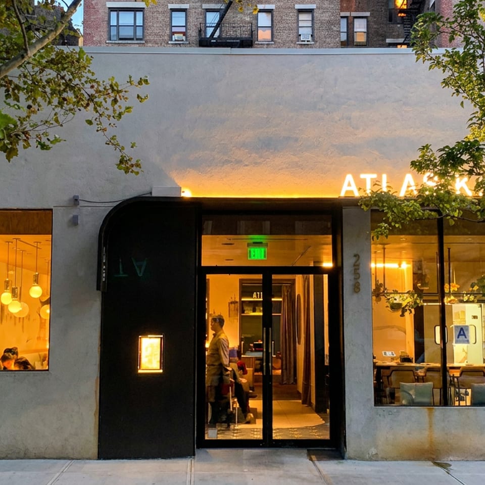 Evening view of a warmly lit Asian kitchen restaurant storefront with glowing signage, large windows, trees, and diners inside.