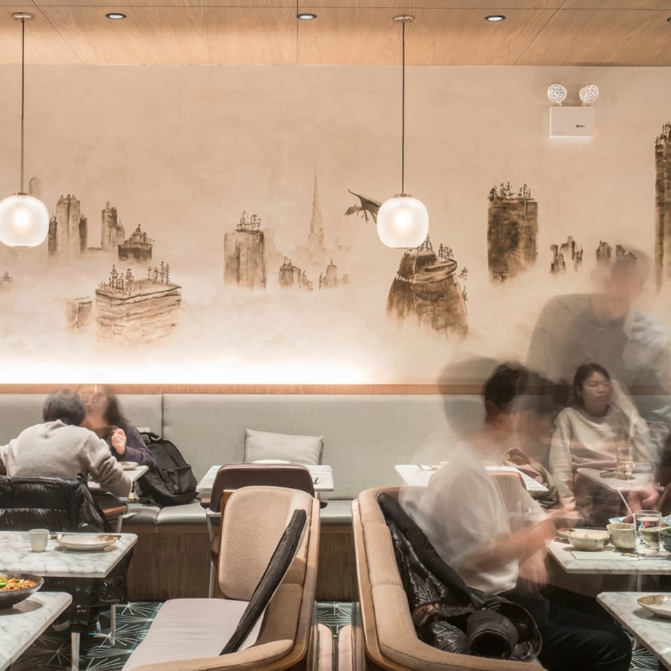 Busy restaurant dining room with groups of diners, plated dishes, pendant lights, and a misty illustrated cityscape mural.