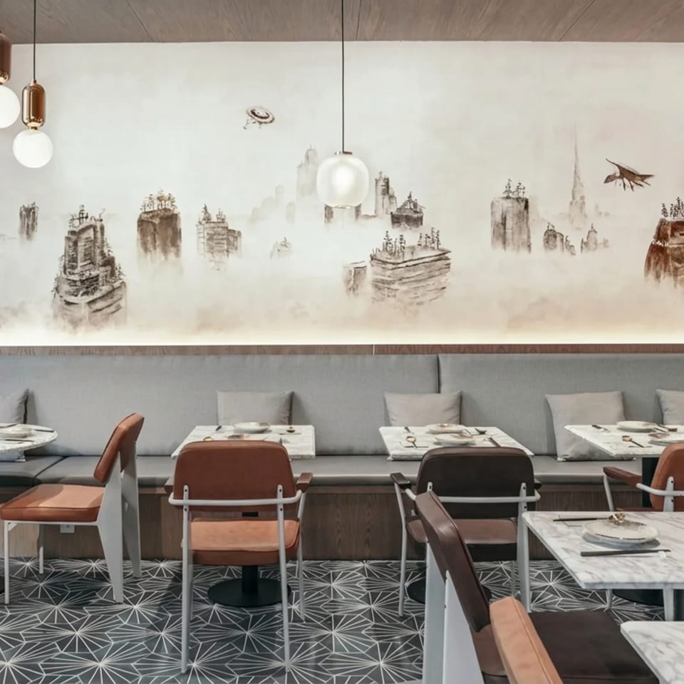 Modern restaurant dining room with set marble tables, upholstered banquette seating, pendant lights, and a misty cityscape mural.