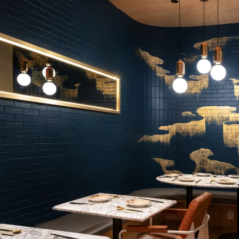 Elegant restaurant dining area with dark blue brick walls, gold artwork, pendant lights, marble tables, and cushioned seating.