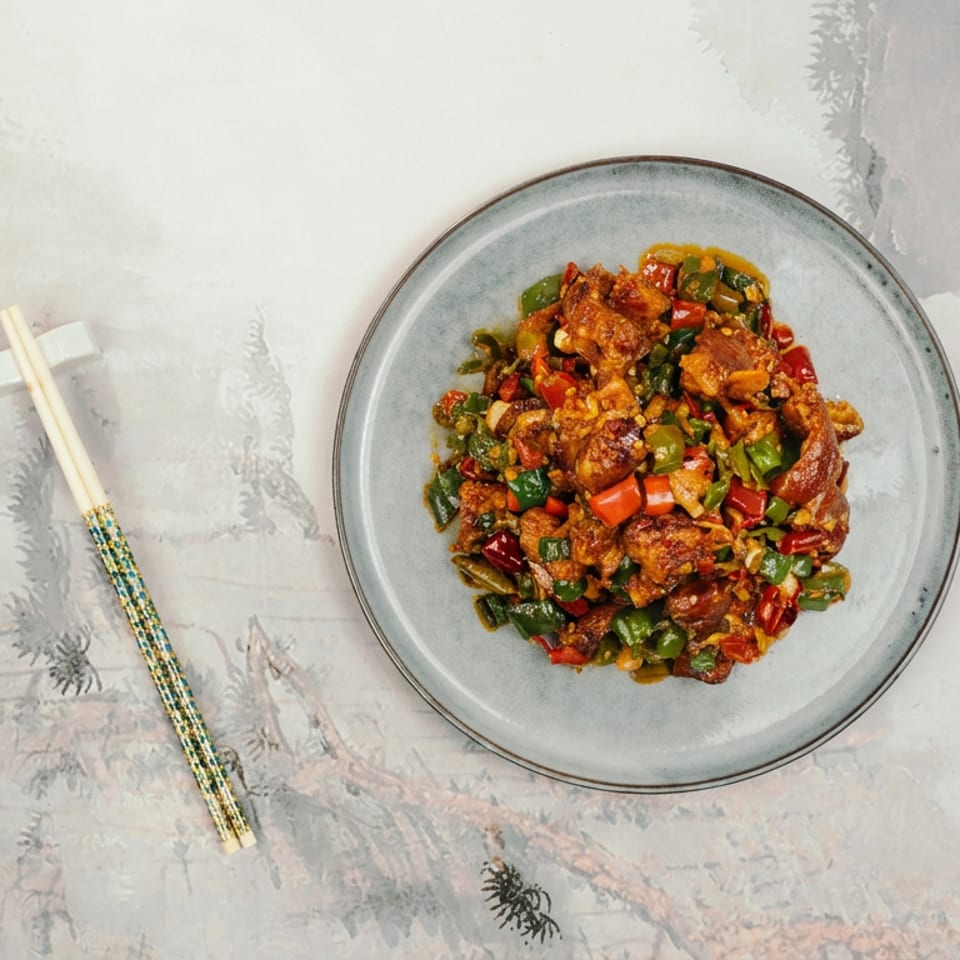 Plate of spicy stir-fried chicken with red and green peppers, served beside patterned chopsticks on a decorative tablecloth.